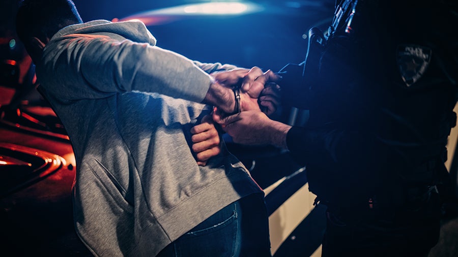 A police officer in a dark uniform putting handcuffs on a person in a grey hoodie, under flashing blue and red lights.