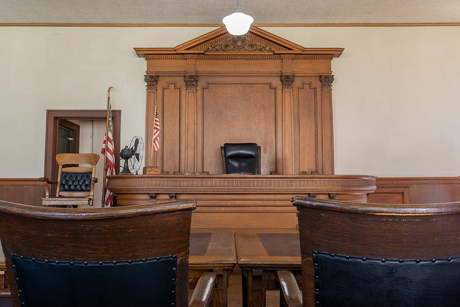 Ornate wooden courtroom with a judge's bench, black leather chair, American flags, and two spectator chairs.
