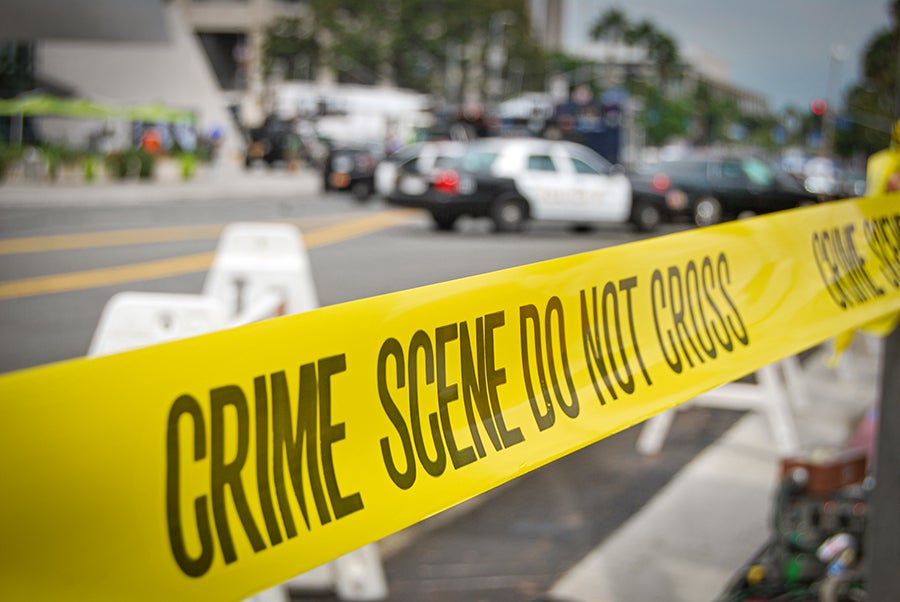 Yellow CRIME SCENE DO NOT CROSS tape blocks a street with blurred police cars and buildings in the background.
