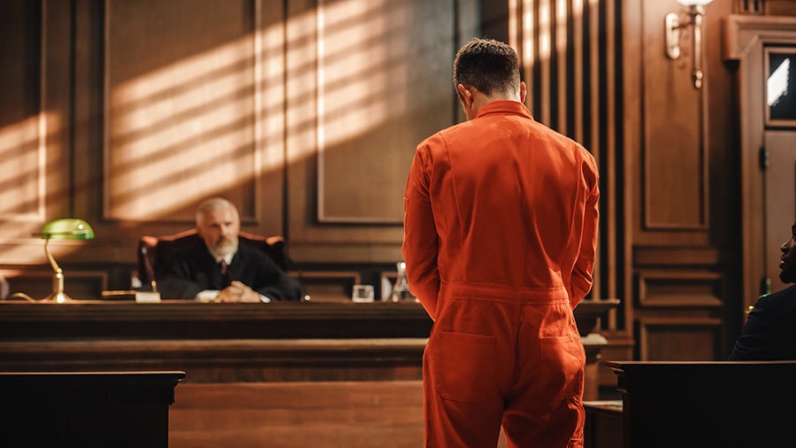 Rear view of a person in an orange jumpsuit facing a judge in a formal courtroom.