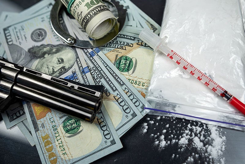A handgun, handcuffs, $100 bills, a bag of white powder, and a syringe on a dark surface.