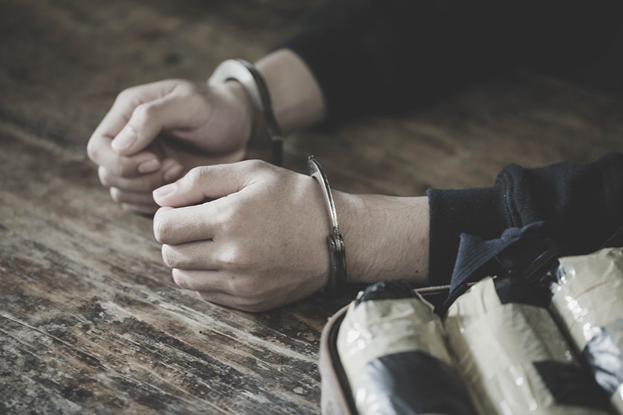 Man's handcuffed hands on a wooden table, next to a bag open with foil-wrapped packages.