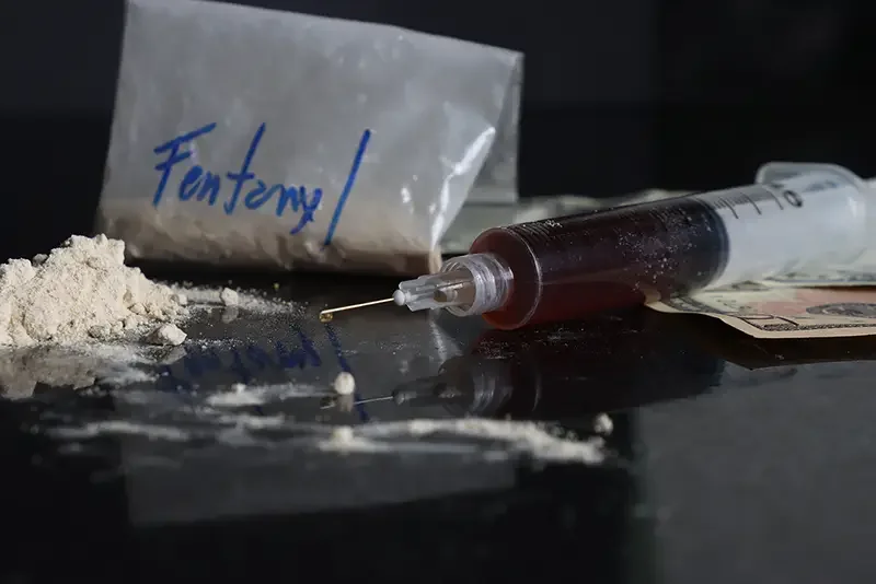 A bag of fentanyl, a syringe with blood, and a pile of white powder on a dark surface, with money in the background.