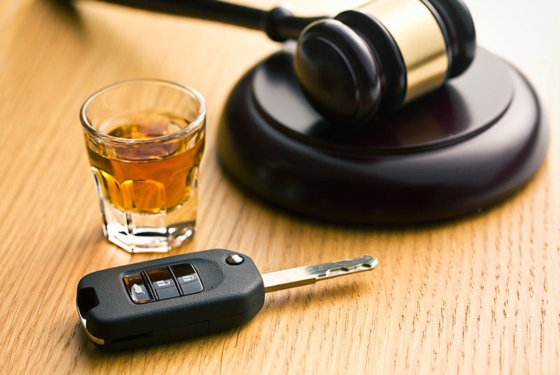 A shot glass of alcohol, car keys, and a judge's gavel on a wooden surface, representing driving under the influence.