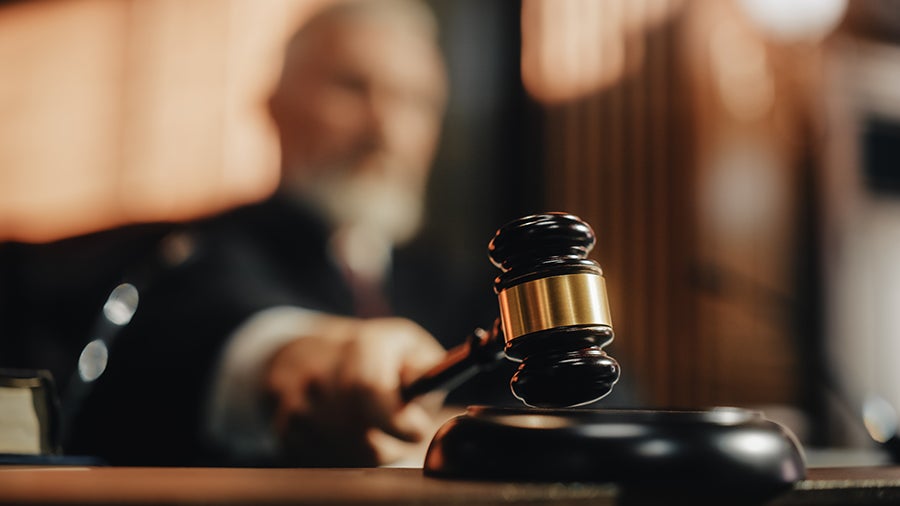 A wooden gavel being struck onto a sounding block on a judicial bench, with a blurred judge in the background.