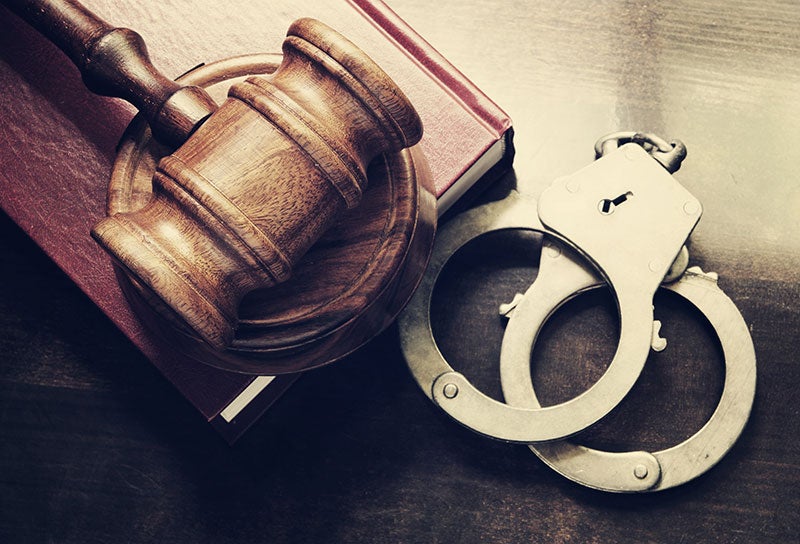 A wooden gavel rests on a red book next to a pair of silver handcuffs, all on a dark wooden surface.