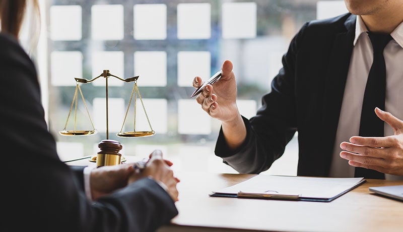A lawyer in a suit and tie discusses a legal document with a client at a desk, with scales of justice nearby.
