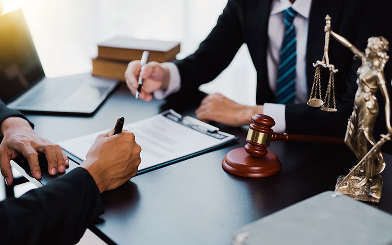 Two people in suits at a desk with legal documents, a judge's gavel, and a statue of Lady Justice.