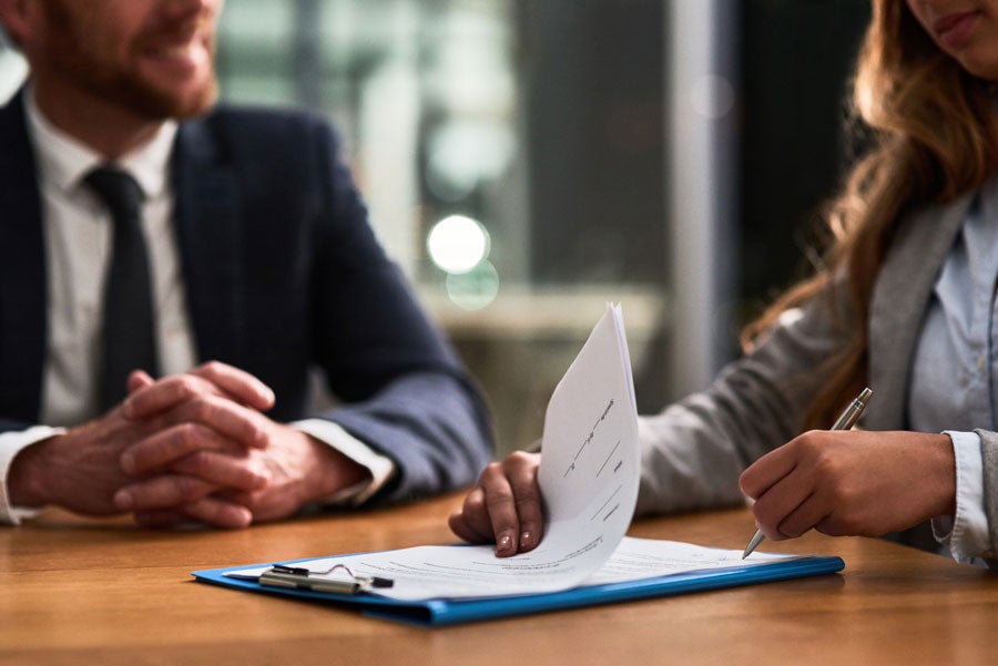 A woman signs a document on a clipboard while a man in a suit watches, sitting across a wooden table.