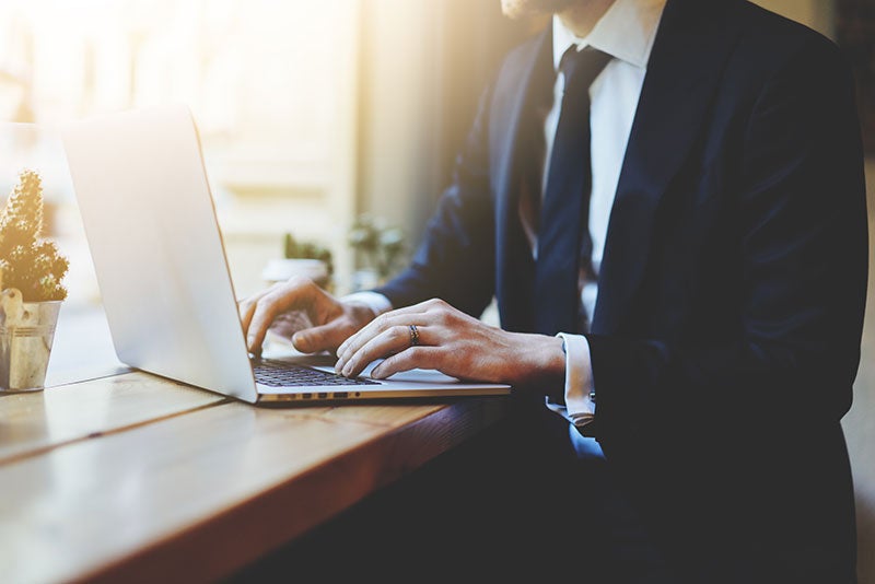 Close-up of a well-dressed person's hands typing on a laptop computer at a light-filled wooden table.