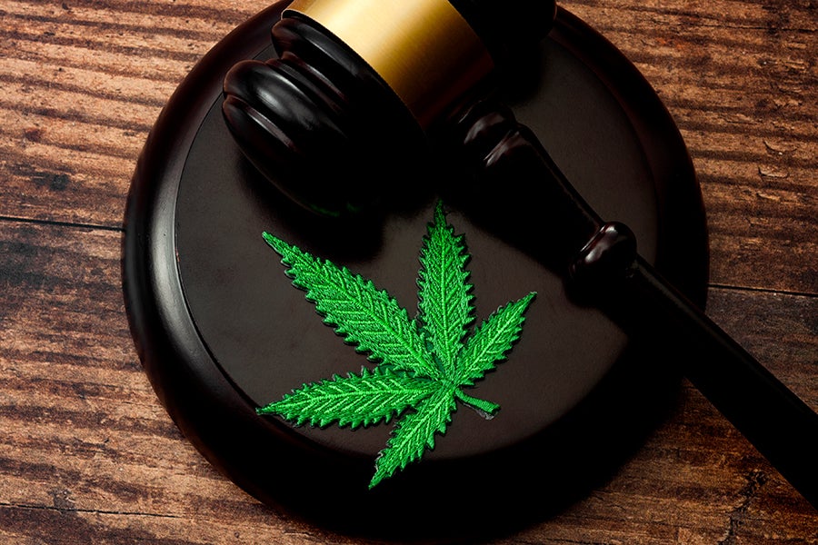 A green embroidered cannabis leaf rests on the black wooden sound block of a judge's gavel on a wooden table.