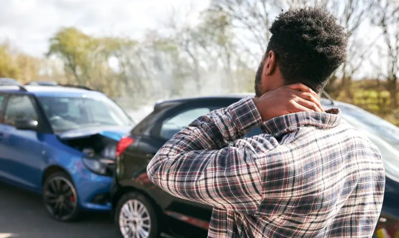 Man clutching his neck after a car accident between two damaged vehicles on a paved road.
