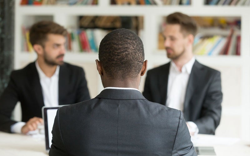 Three men in suits in an office; one from the back, two facing him, blurred. One man has a laptop.