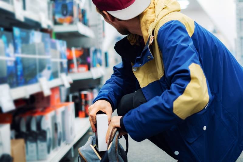 A person in a blue and yellow jacket shoplifts an item from a store display, putting it into a bag.