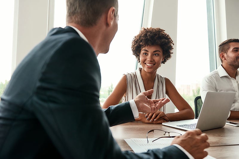 A smiling Black woman listens intently to a man in a business suit, who gestures with his hand during a meeting.