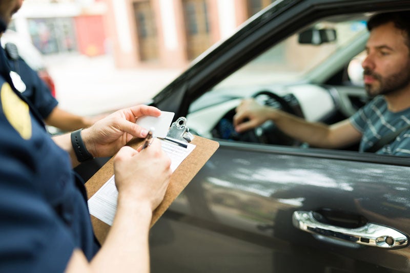 A police officer writing a ticket on a clipboard, while a man in a car looks on from the driver's seat.