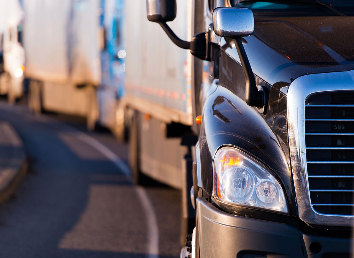 Close-up of a black semi-truck's front corner on a road, with more semi-trucks blurred in the background.