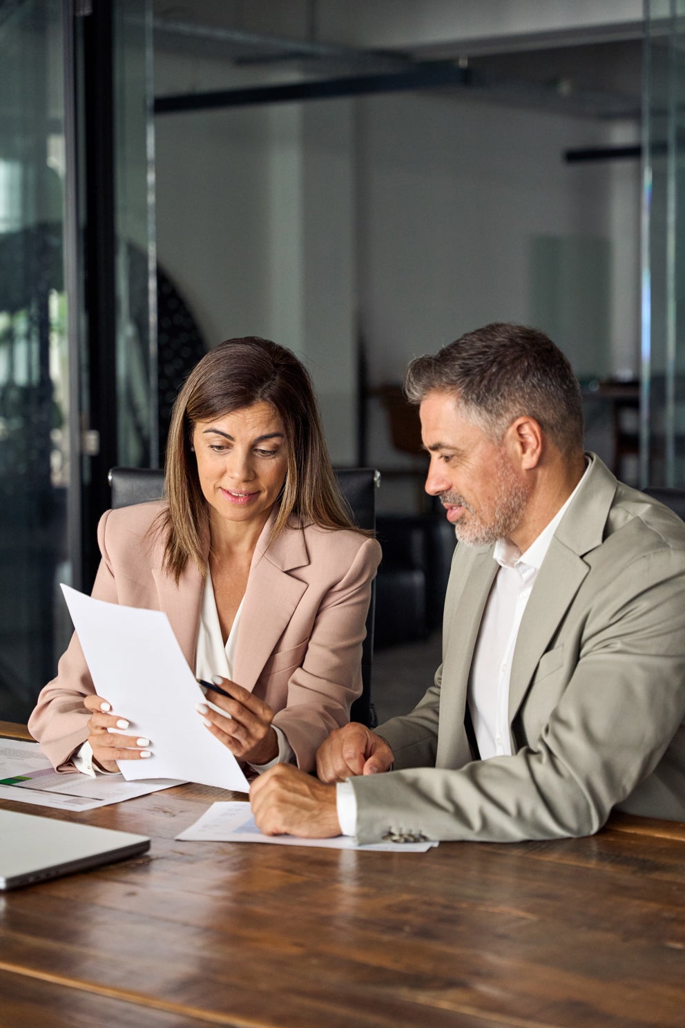 Two professionals, a woman and a man, review documents together at a dark wooden table in an office setting.