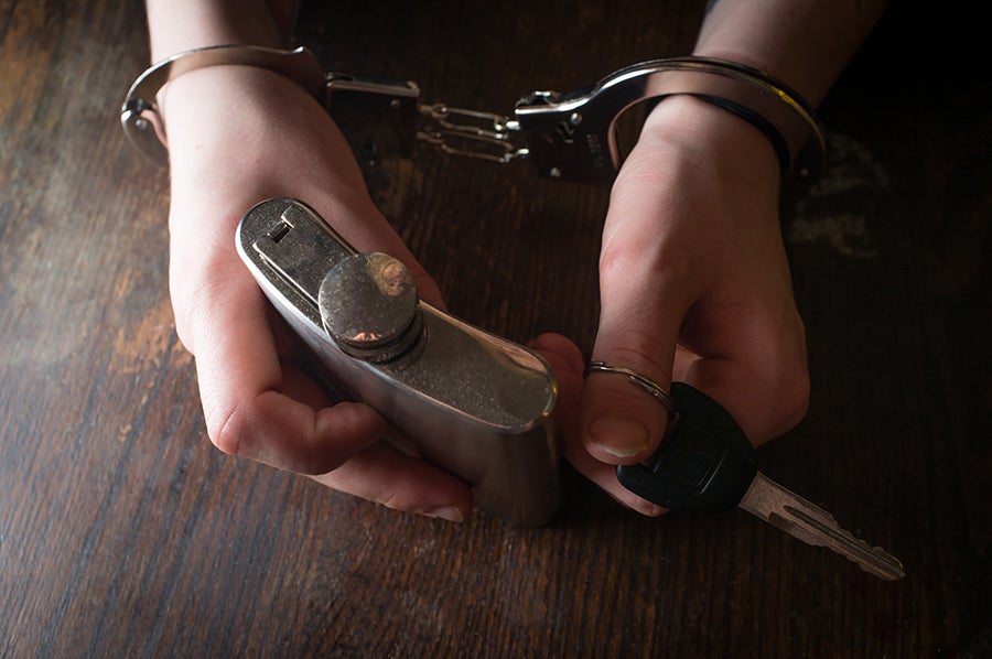 Hands in handcuffs hold a flask on the left and car keys on the right, on a dark wooden table.