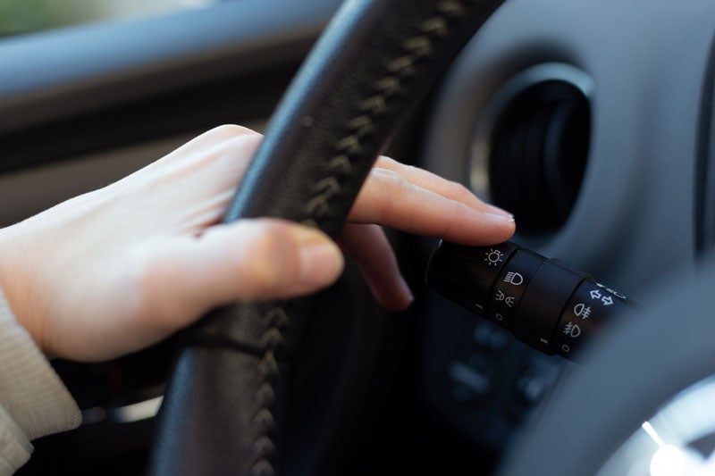 A person's hand on a car's steering wheel, pressing their finger on the light switch symbol on the indicator stalk.