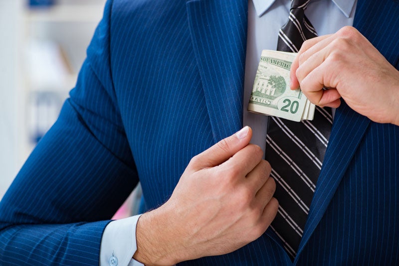 A man in a blue pinstripe suit and striped tie putting a wad of twenty-dollar bills into his inner jacket pocket.